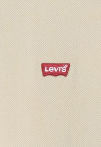 Etiqueta de tela roja con el texto "Levi's" en blanco sobre un fondo beige texturizado. La etiqueta tiene una forma distintiva curva en los bordes superiores.