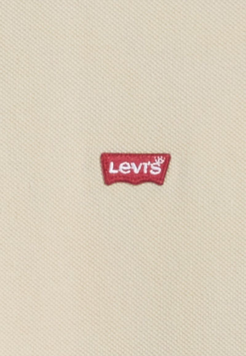 Etiqueta de tela roja con el texto "Levi's" en blanco sobre un fondo beige texturizado. La etiqueta tiene una forma distintiva curva en los bordes superiores.