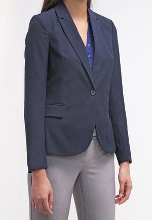 Donna che indossa una giacca blu scuro aderente sopra una camicia blu con bottoni e pantaloni grigio chiaro, in piedi davanti a uno sfondo chiaro e semplice.