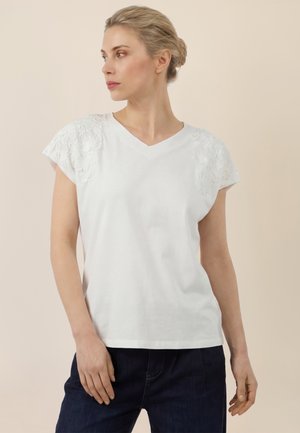 Frau mit blondem Haar zu einem Dutt gebunden, trägt ein weißes V-Ausschnitt-Oberteil mit Spitzen an den Schultern und dunkelblaue Jeans, blickt nach links vor beigem Hintergrund.