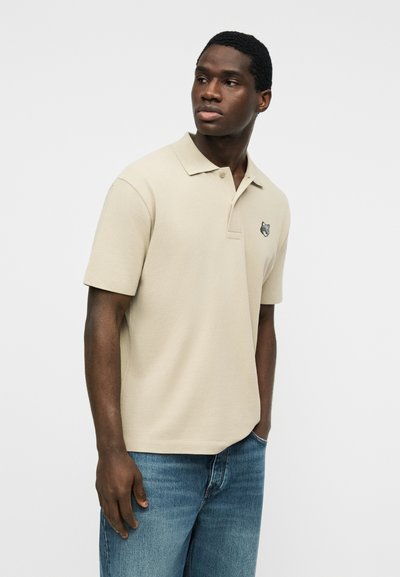 Άνδρας με κοντομάνικο μπέζ πόλο μπλουζάκι με λογότυπο λύκου στο στήθος, φοράει μπλε τζιν και κοιτάζει προς το πλάι μπροστά σε έναν απλό φόντο.