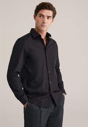 Chemise noire boutonnée avec col classique, manches longues et tissu en coton lisse ; associée à un pantalon gris. Présente des détails de couture subtils.
