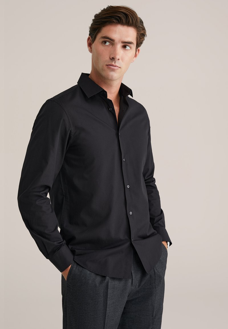 Camisa negra abotonada con cuello clásico, mangas largas y tela suave de algodón; combinada con pantalones grises. Presenta detalles sutiles de costura.