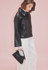 Čierna syntetická kožená bunda s golierom, prednými gombíkmi a dvoma vreckami na hrudi, skombinovaná s čiernou kabelkou a bielymi nohavicami.