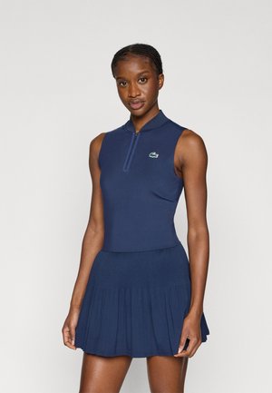 Marineblauwe mouwloze jurk met een hoge ritssluiting bij de kraag, een aangesloten lijfje en een uitlopende rok. Voorzien van een klein groen logo op de borst. Glad oppervlak.