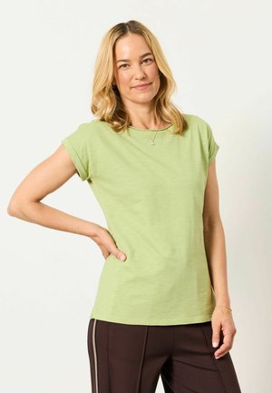 Blonde woman wearing a light green short-sleeve shirt and dark pants, standing with one hand on her hip, smiling gently.