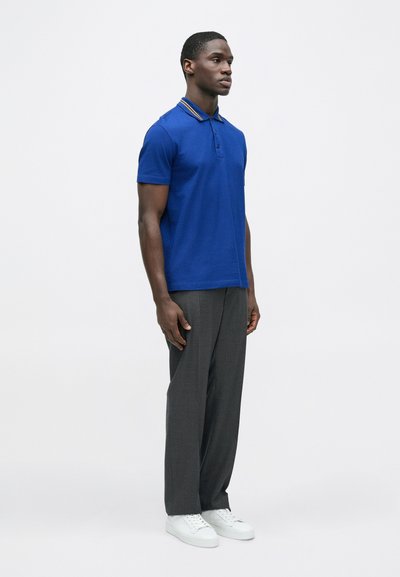 Vyrų mėlynas polo marškinėliai su trumpomis rankovėmis, kontrastingomis apykaklės juostelėmis, derinami su pilkomis kelnėmis ir baltais sportbačiais. Paprastas, klasikinio dizaino.