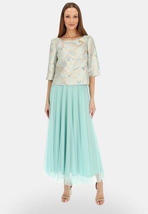 Kobieta nosząca jasnoniebieski kwiecisty top z brokatu z rękawami do połowy przedramienia i dopasowaną długą plisowaną tiulową spódnicę, pozująca z rękami wzdłuż ciała.