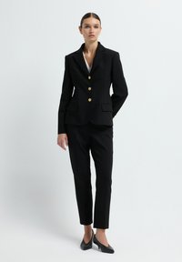 Zwarte op maat gemaakte pak met een getailleerde blazer met gouden knopen, twee voorzakken en slanke broeken. Het materiaal heeft een soepele textuur.