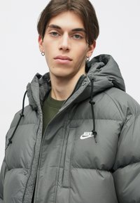 Casaco cinza isolado com capuz, apresentando um fecho frontal, detalhes em cordão e um pequeno logótipo branco da Nike no peito.