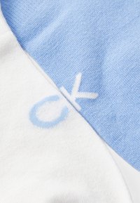 Artimųjų planų švelnaus mezginio audinys šviesiai mėlynos ir baltos spalvos, ant kiekvienos spalvos dalies matomos dalinės raidės „C“ ir „K“.