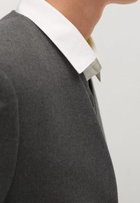 Personne portant un blazer gris foncé sur une chemise blanche à col, montrée du cou à la partie supérieure de la poitrine sur un fond uni.