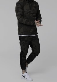 Person iført mørk camouflagejakke og bukser med hvide Nike sneakers, stående på en ensfarvet lys-grå baggrund.