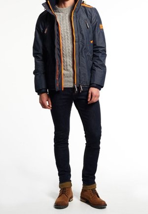 Granatowa kurtka puchowa z pomarańczowymi akcentami, kieszeniami na zamek i wysokim kołnierzem, noszona na jasnoszarym swetrze w warkocze i ciemnych dżinsach. Brązowe buty.