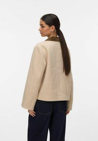 Veste beige en mélange de laine avec un design court, des manches larges et un col marron foncé, associée à un jean taille haute bleu foncé.