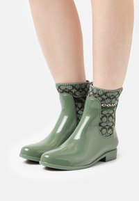 Botas de tobillo de goma verde con un calcetín tejido con patrón, que presenta un diseño verde oscuro y el logotipo de la marca, acabado liso y brillante, y un pequeño tacón cuadrado.
