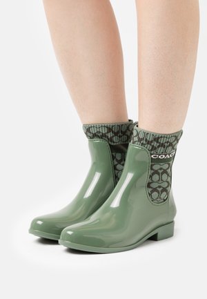 Botas de tobillo de goma verde con un calcetín tejido con patrón, que presenta un diseño verde oscuro y el logotipo de la marca, acabado liso y brillante, y un pequeño tacón cuadrado.