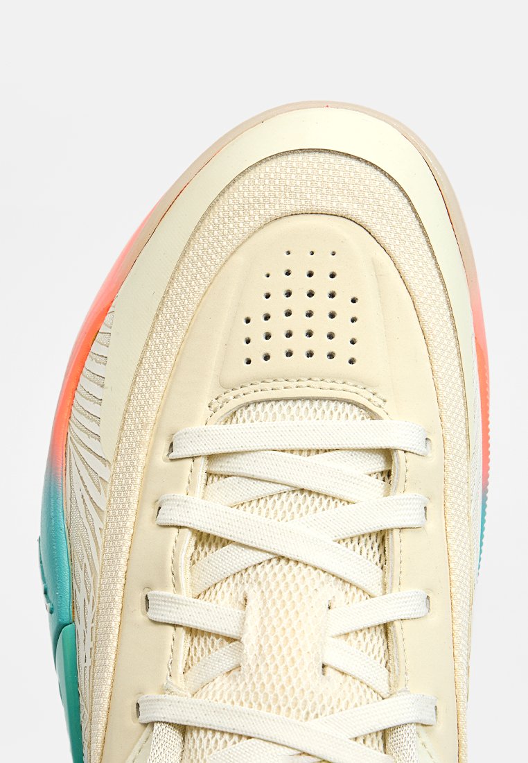 Zapatilla con parte superior beige que presenta texturas de malla y cuero, cordones blancos y una suela degradada en tonos de naranja a turquesa.