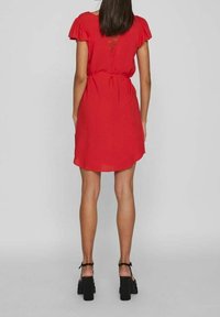 Vestido corto rojo con mangas acampanadas, cintura ajustada y dobladillo curvado. Presenta un detalle de encaje en la parte posterior y se combina con zapatos de tacón negros con tiras.