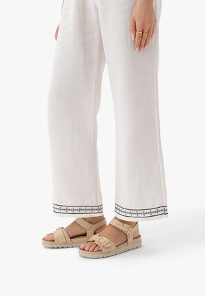 Kobieta ubrana w białe spodnie z szerokimi nogawkami z czarną haftowaną dekoracją na mankietach oraz beżowe sandały z klamrą na białym tle.