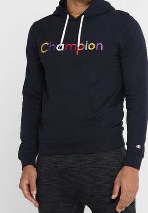 Person iført en sort hættetrøje med flerfarvet "Champion"-logo på brystet og hvide snore, kombineret med mørkegrå bukser.