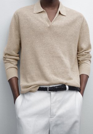 Beiger Strickpullover mit langen Ärmeln und offenem Kragen, in weiße Hose gesteckt, getragen mit einem schwarzen geflochtenen Gürtel, Hände in den Taschen.