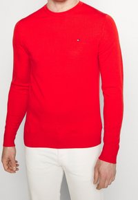Czerwony sweter z okrągłym dekoltem i długimi rękawami, wykonany z gładkiego materiału dzianinowego. Ma małe logo na piersi. Noszony z białymi spodniami.