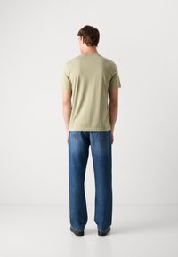 Olivenfarget t-skjorte med korte ermer, kombinert med løse blå denimjeans, sett fra bak på en ensfarget bakgrunn.