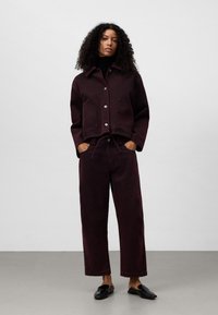 Kvinna med lockigt hår som bär en mörklila knappad jacka, matchande vida byxor, svart turtleneck och svarta slip-on skor.