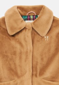 Chaqueta de piel sintética color tostado con un amplio collar, cierre de un botón y un alfiler decorativo en forma de lazo dorado. Forrada con una tela de cuadro de colores.