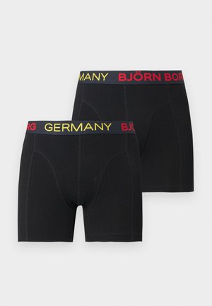 Atletski tamnoputi muškarac stoji bos, nosi crne bokserice s natpisom "NJEMAČKA" i "BJÖRN" na pojasu, gleda u stranu.