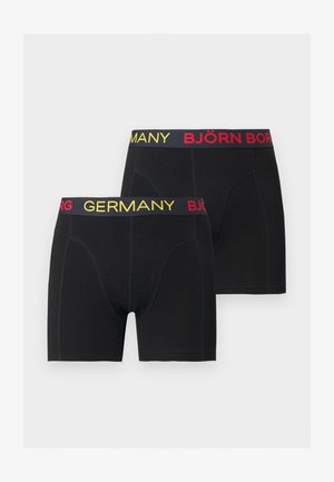 Homme athlétique à la peau foncée debout pieds nus, portant un boxer noir avec "ALLEMAGNE" et "BJÖRN" sur la ceinture, regardant sur le côté.