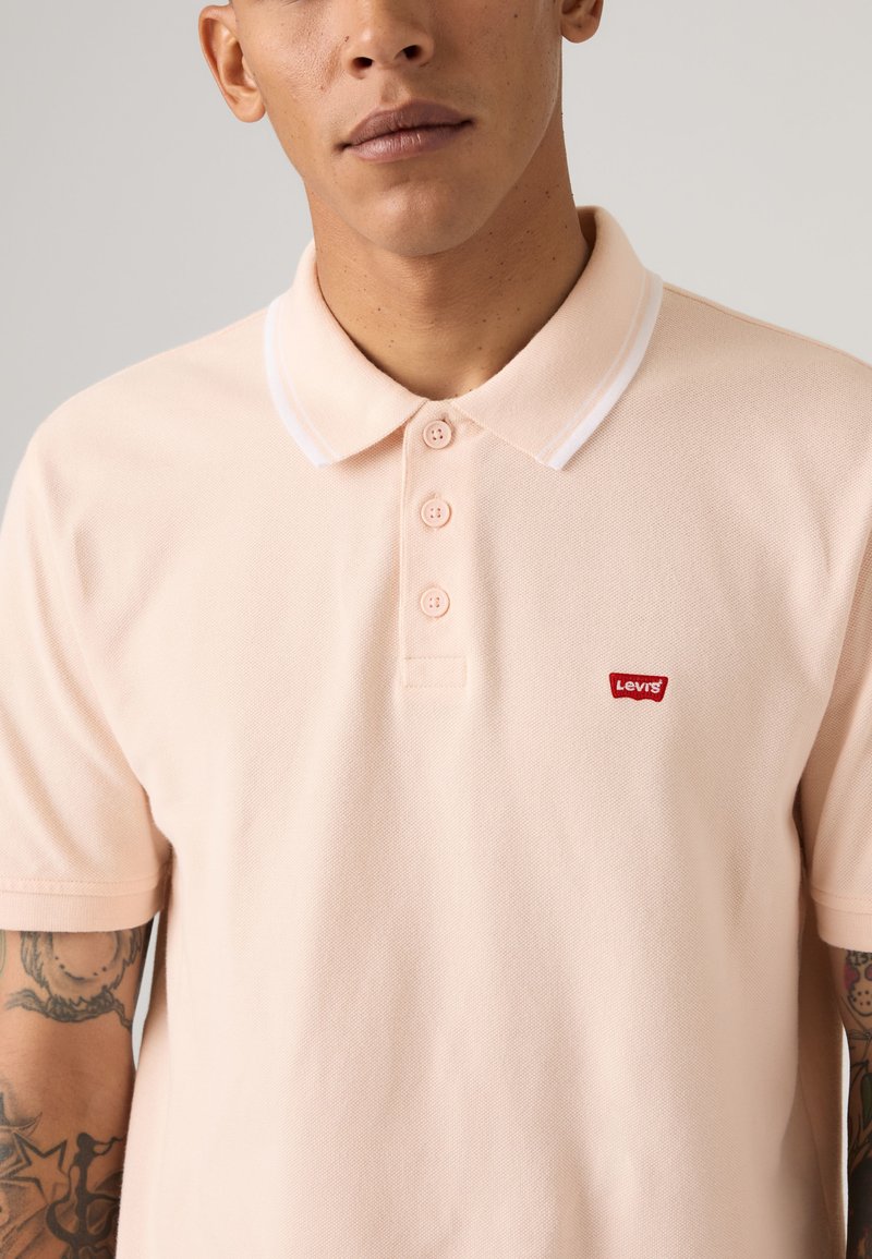Homme portant un polo Levi's rose clair avec trois boutons et des tatouages visibles sur les deux avant-bras, sur fond neutre.
