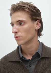 Pull en maille marron sur une chemise grise à col, mettant en valeur un contraste de tissus lisses. Le mannequin a les cheveux courts et décoiffés et une texture de peau naturelle.