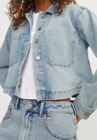 Svetle modrá džínsová bunda s cropped designem, stříbrnými knoflíky, dvěma předními kapsami a detaily šití. Nositá přes bílý top.