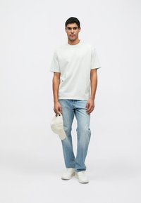 Világosszürke, kerek nyakú póló, kék farmernadrág és fehér tornacipő. Világos bézs sapkát tart a kezében. Egyszerű dizájn, hétköznapi szabás.