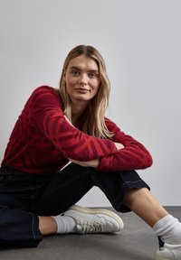 Rot gemusterter Pullover mit weicher Textur, dunkelblaue, verkürzte Jeanshosen und weiße Sneaker mit grauen Akzenten. Modell sitzt vor einem neutralen Hintergrund.