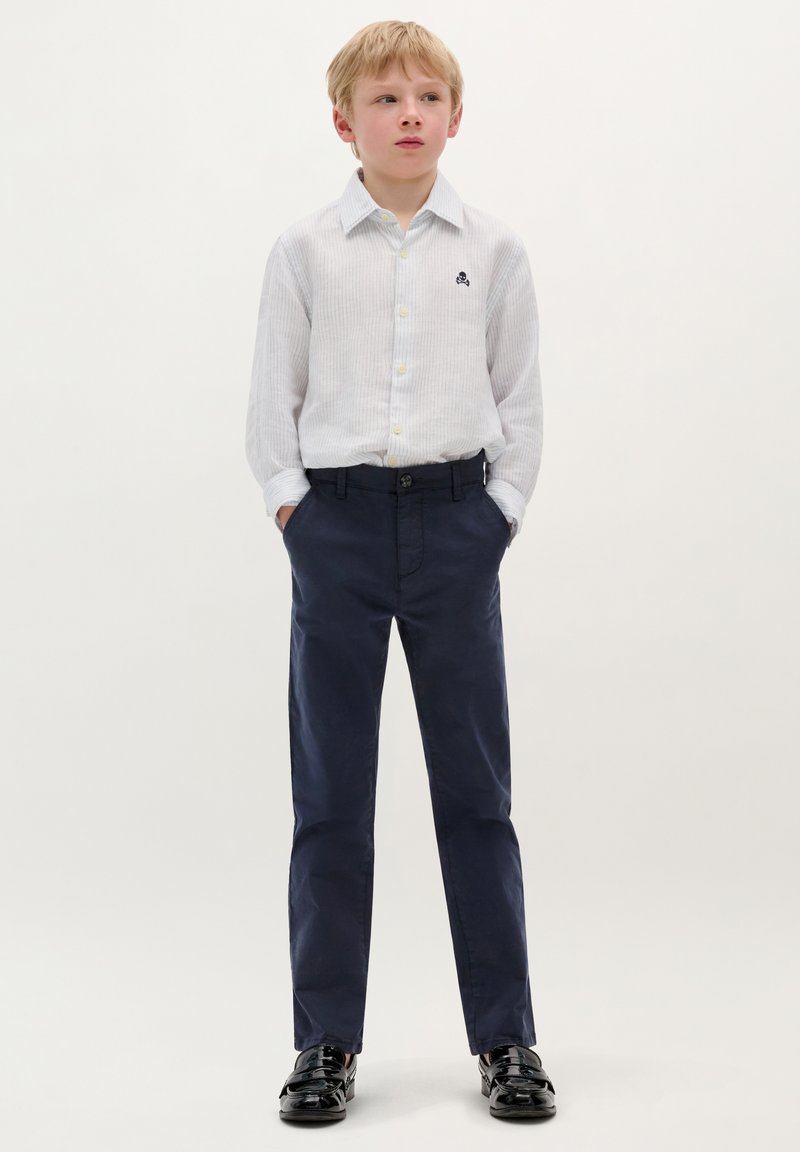 Cămașă cu dungi albe și nasturi galbeni; pantaloni bleumarin; pantofi negri; design simplu cu detalii logo pe cămașă.