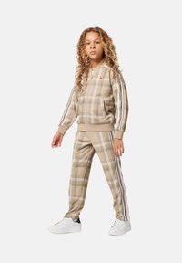 Enfant avec de longs cheveux bouclés portant un survêtement à carreaux beige avec des détails en rayures blanches et des baskets blanches, se tenant devant un fond uni.