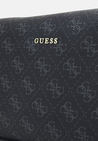 Sac à main noir texturé avec un motif en losange et un logo embossé, accentué par le nom de marque doré "GUESS" sur le devant.