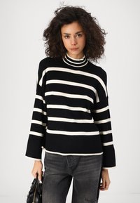 Črno-bela striped pletena pulover z visokim ovratnikom, dolgimi zvončastimi rokavi in rebrasto teksturo. Kombiniran s temno sivo uničenimi jeans hlačami.