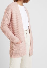 Hellrosa gestrickte cardigan mit strukturiertem Muster, offener Vorderseite und zwei Seitentaschen. Lange Ärmel mit gerippten Manschetten.