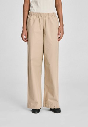 Isik kannab beežikaid laia säärega pükse elastse vöökohaga, musti baleriinakingi ja valget varrukateta soonikkoes plussi.