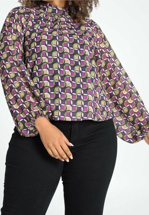 Kvinde iført en lilla, grøn og beige bluse med geometrisk mønster og lange ærmer samt sorte bukser, der viser overkrop og hånd.