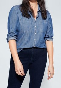 Jasnoniebieska koszula z dżinsu z zapięciem na guziki i podwiniętymi rękawami, z dwoma kieszeniami na piersiach i kontrastującym szwem, zestawiona z ciemnoniebieskimi spodniami.