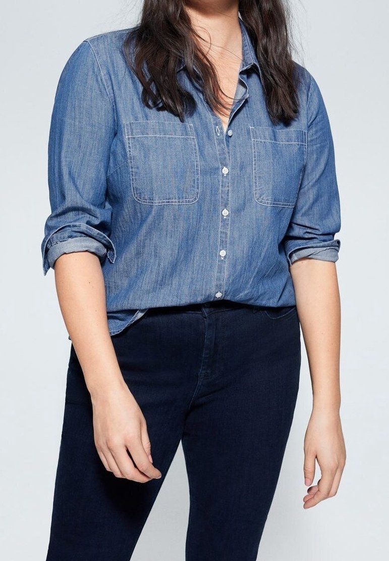 Jasnoniebieska koszula z dżinsu z zapięciem na guziki i podwiniętymi rękawami, z dwoma kieszeniami na piersiach i kontrastującym szwem, zestawiona z ciemnoniebieskimi spodniami.