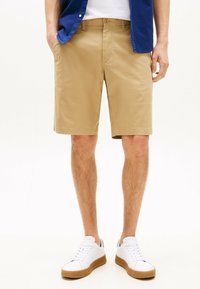Shorts beiges avec une texture lisse, coupe droite, deux poches latérales, portés avec des baskets blanches à semelles en caoutchouc et une chemise bleue.