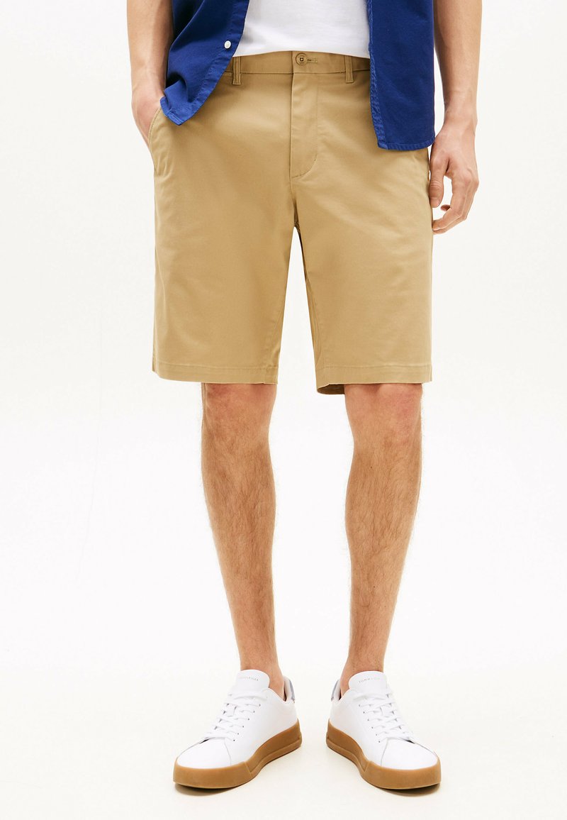 Shorts beiges avec une texture lisse, coupe droite, deux poches latérales, portés avec des baskets blanches à semelles en caoutchouc et une chemise bleue.