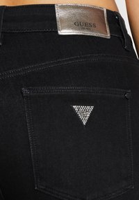 Jeans de denim negro con una etiqueta metálica plateada "GUESS" en la cintura y un triángulo decorativo de strass en el bolsillo trasero.