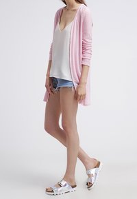 Lång rosa kofta i lätt material, tillsammans med en vit topp och slitna jeanstrosor. Skorna är blanka silverfärgade slidesandaler.